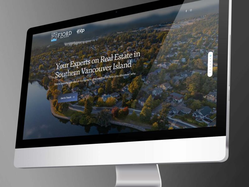 A computer monitor angled in 3D displaying a real estate website hero for Southern Vancouver Island with an aerial neighborhood view.